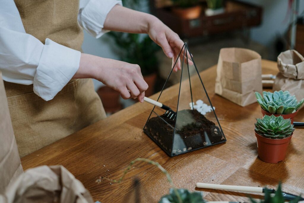 Hands crafting a succulent terrarium with a triangular glass prism indoors.