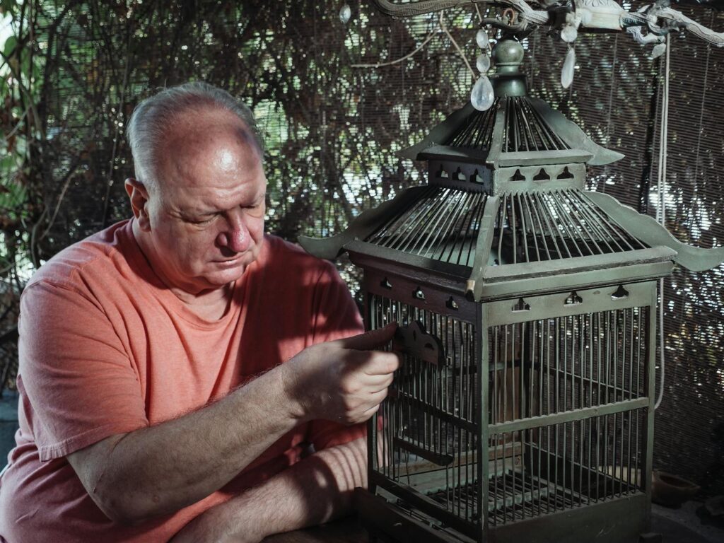 Elderly man in contemplation, sitting indoors by an empty birdcage. Themes of reflection and solitude.
