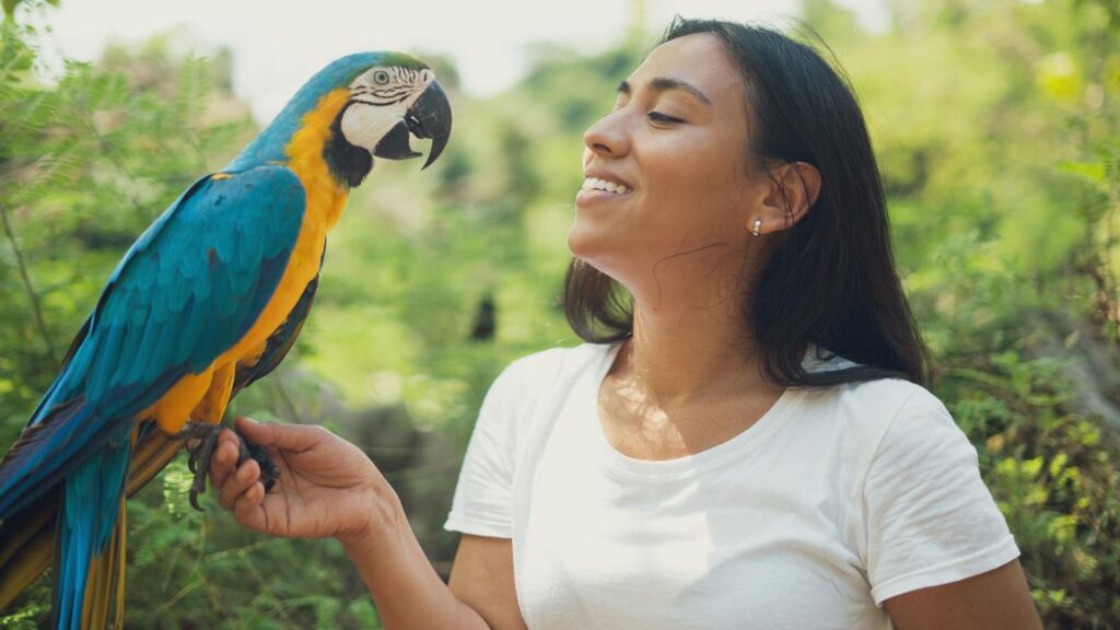 A woman lovingly interacts with a blue and gold macaw in a lush outdoor setting.
