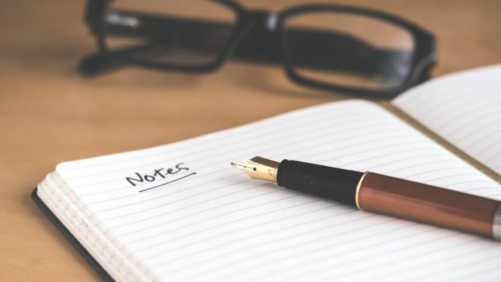 Open notebook with handwritten "Notes," a fountain pen, and glasses on a desk.