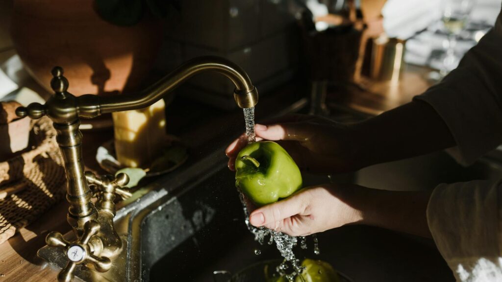 Hands washing fresh green bell pepper under a vintage faucet in a warm, rustic kitchen setting.