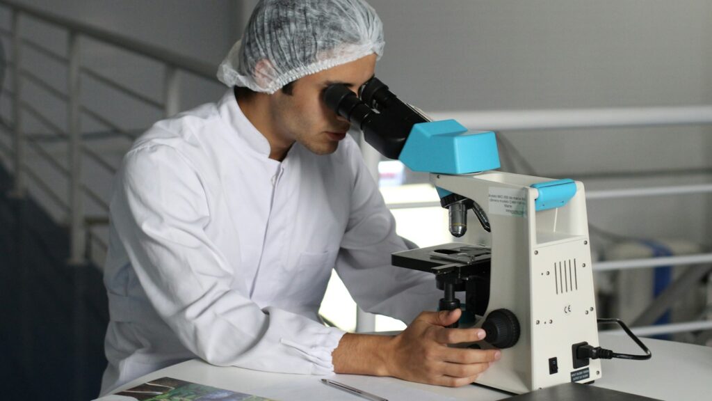 Scientist in a lab coat and hairnet using a microscope for research or analysis.