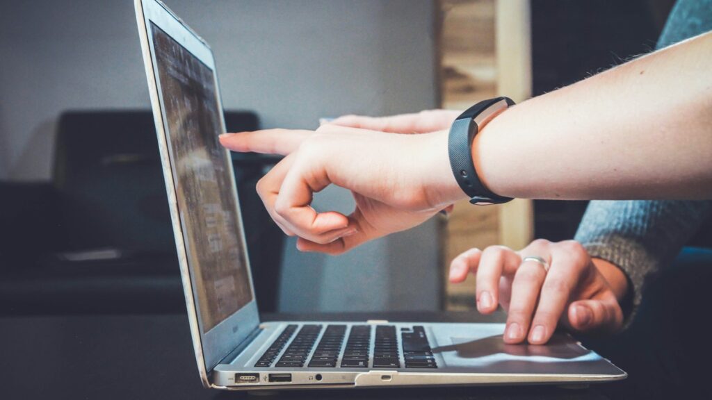 Two hands are shown interacting with a laptop; one finger points to the screen while the other hand is on the keyboard.