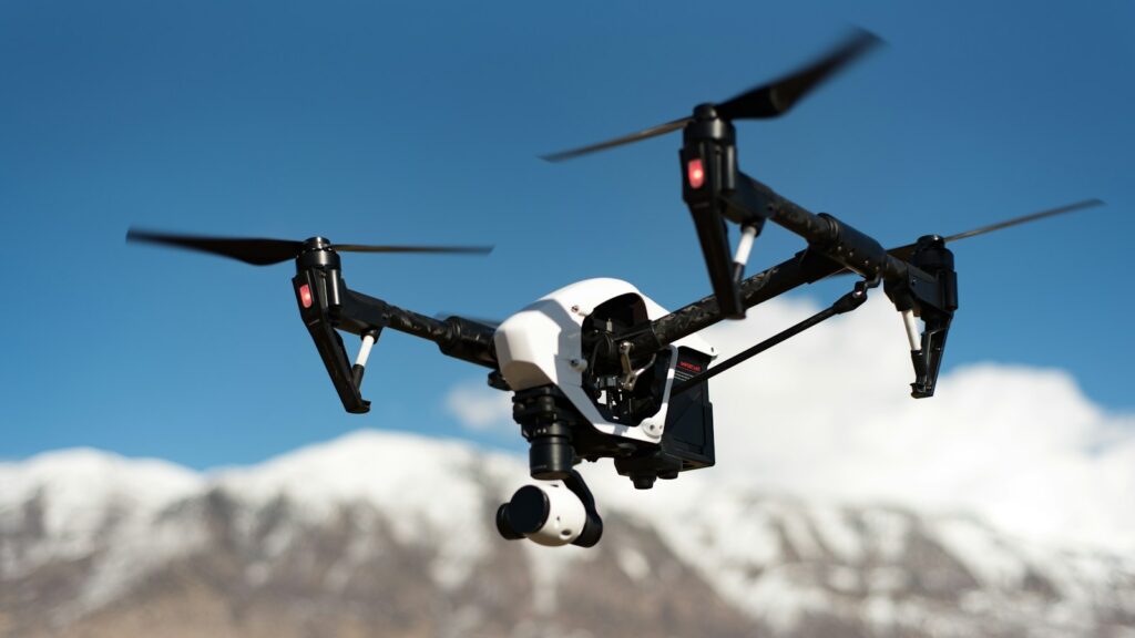 A black and white quadcopter drone hovering under the blue sky with snowy mountains in the background.